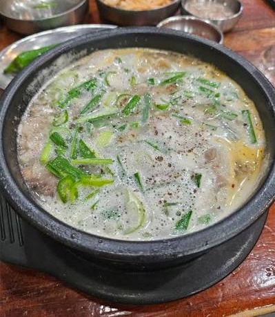 A bowl of suyuk-gukbap in a black earthenware pot: milky pork broth with slices of boiled pork under the surface, topped with chopped scallions and ground pepper, banchan dishes behind it on a wooden table