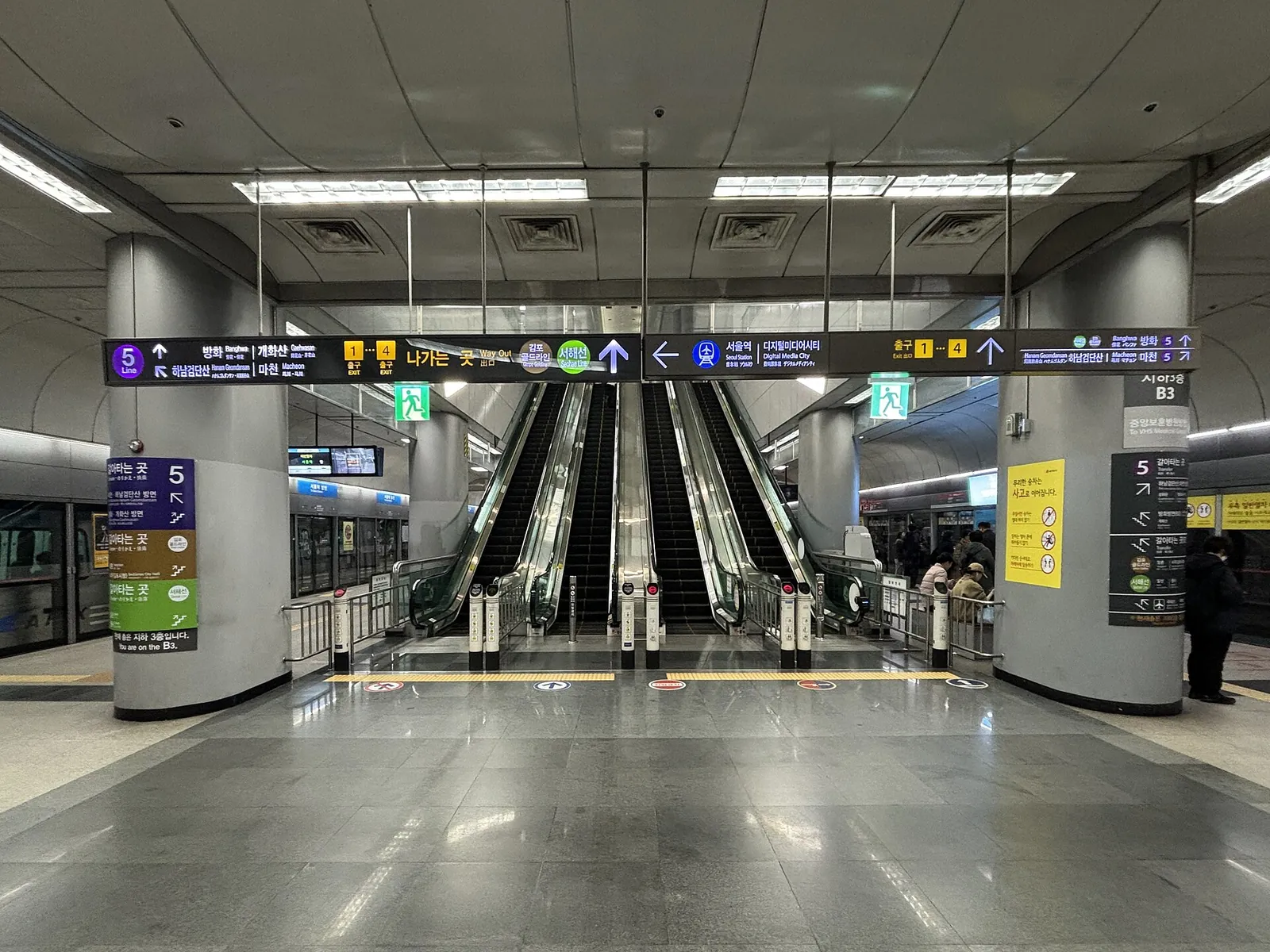 Underground platform at Gimpo Airport station with escalators in the middle, a train behind platform screen doors on the left, and overhead signs pointing to Line 5, Seoul Station, Digital Media City and the exits