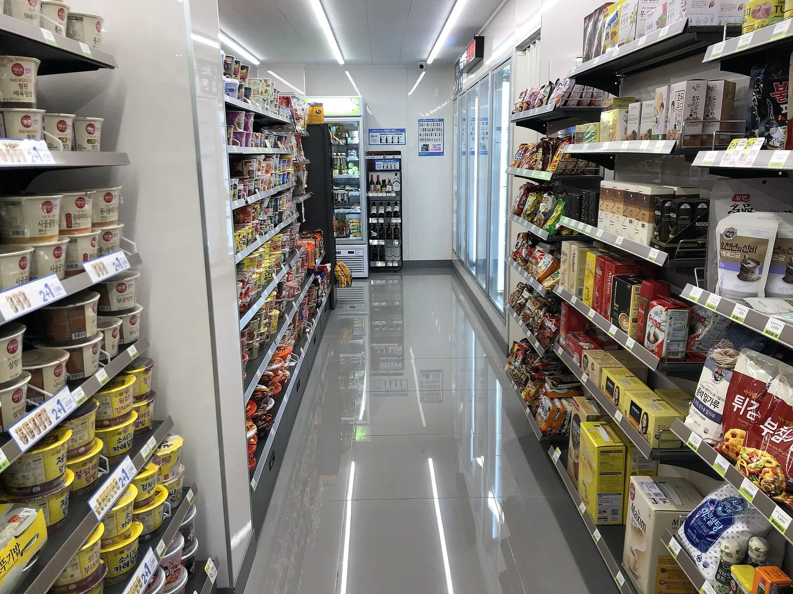 Narrow aisle inside a CU convenience store with shelves of instant rice bowls on the left marked with blue 2+1 shelf tags, cup noodles further down, and drinks fridges along the right wall