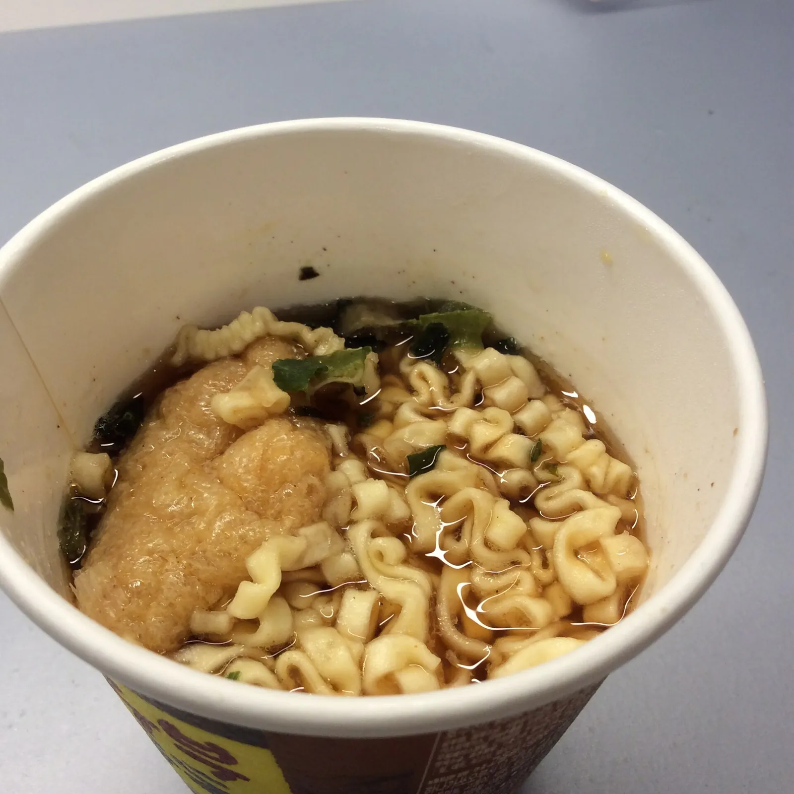 Top-down view into a paper cup of instant ramyeon after hot water was added: wavy noodles, a piece of fried tofu and green onion flakes in brown broth