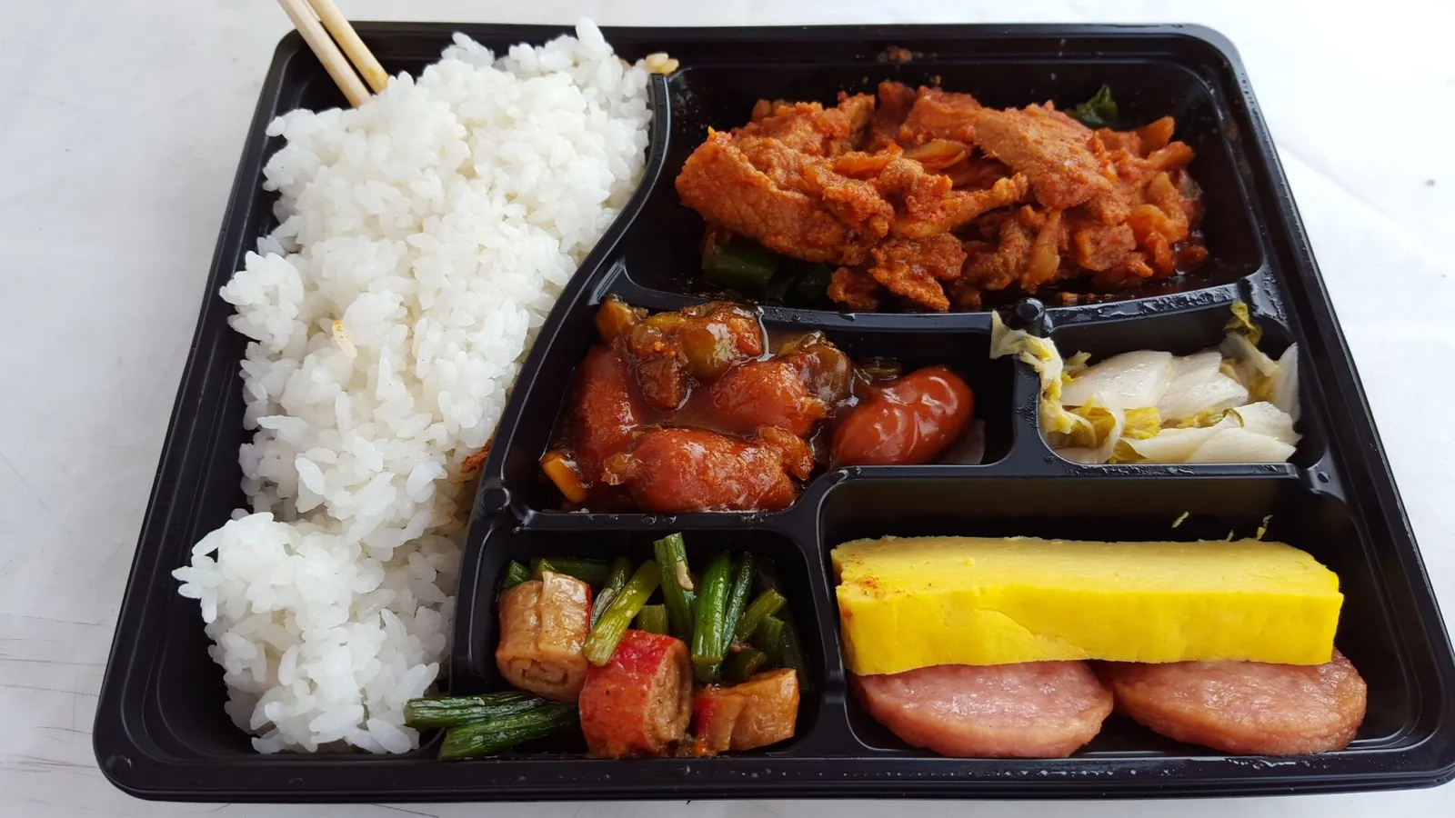 A Korean convenience store lunchbox in a black plastic tray: white rice, spicy stir-fried pork, sausages in sauce, napa cabbage kimchi, green beans with fish cake, a slab of rolled omelet and two slices of ham