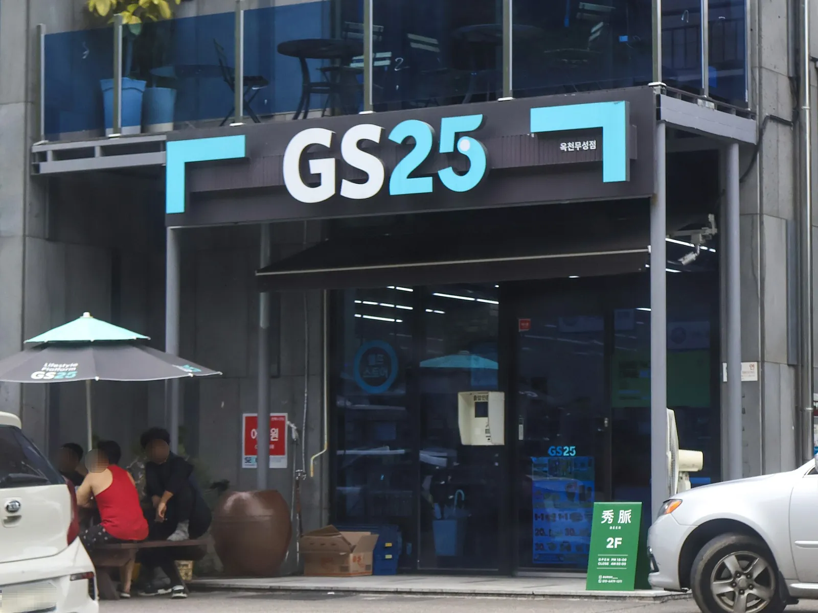 Front of a GS25 convenience store with the GS25 sign over a glass entrance, a patio umbrella and an outdoor table beside the door
